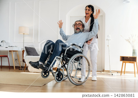 Elderly man in wheelchair smiles with arms raised while caregiver stands behind him in a bright, modern living room with wooden flooring and minimalistic decor Elderly man in wheelchair smiles with arms raised while caregiver stands behind him in a bright, modern living room with wooden flooring and minimalistic decor 142863081
