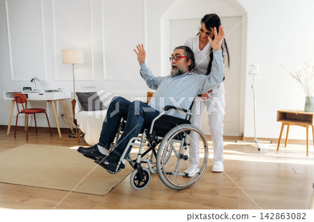Female caregiver assisting elderly man in wheelchair, both smiling, inside a bright, modern living room with wooden flooring and minimalistic decor Female caregiver assisting elderly man in wheelchair, both smiling, inside a bright, modern living room with wooden flooring and minimalistic decor 142863082