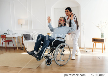 Elderly man in wheelchair joyfully interacts with caregiver in modern living room, bright lighting and minimalist decor create a welcoming atmosphere Elderly man in wheelchair joyfully interacts with caregiver in modern living room, bright lighting and minimalist decor create a welcoming atmosphere 142863083