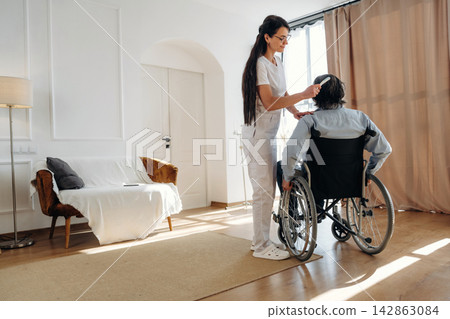 Female caregiver in white uniform assists elderly man in wheelchair inside a well-lit living room with a sofa and curtains, promoting comfort and support Female caregiver in white uniform assists elderly man in wheelchair inside a well-lit living room with a sofa and curtains, promoting comfort and support 142863084