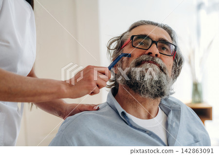 Male client with gray beard and glasses receiving grooming treatment from a beautician in a modern salon setting with soft lighting and minimalistic decor 142863091