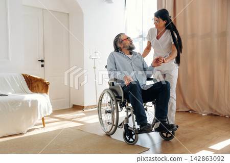 Female caregiver assists elderly man in wheelchair inside a bright, modern living room with wooden flooring and large windows providing natural light Female caregiver assists elderly man in wheelchair inside a bright, modern living room with wooden flooring and large windows providing natural light 142863092