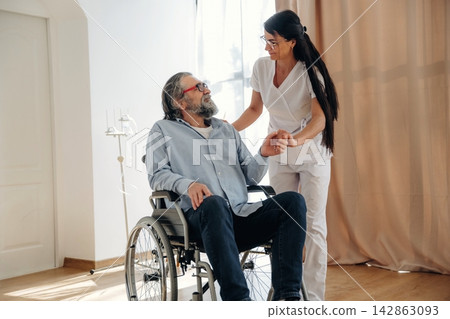 Female caregiver assists elderly man in wheelchair during a health check in a bright room with large windows and soft curtains, promoting comfort and care 142863093