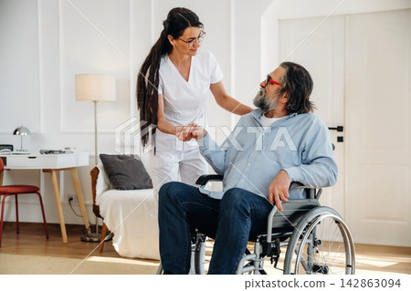 Female caregiver in white uniform assists elderly man in wheelchair inside a bright room with modern furniture and soft lighting, creating a supportive environment 142863094