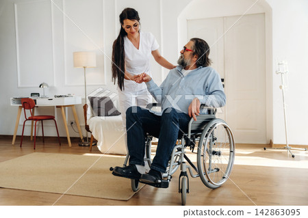 Female caregiver in white uniform assists elderly man in wheelchair inside a bright, modern living room with wooden flooring and minimalistic decor Female caregiver in white uniform assists elderly man in wheelchair inside a bright, modern living room with wooden flooring and minimalistic decor 142863095