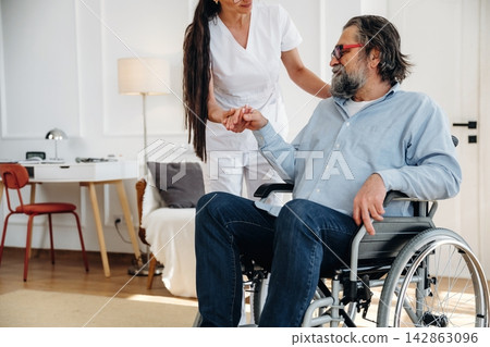 Female caregiver assisting elderly man in wheelchair inside a well-lit room with modern decor, featuring a desk and chair in the background Female caregiver assisting elderly man in wheelchair inside a well-lit room with modern decor, featuring a desk and chair in the background 142863096