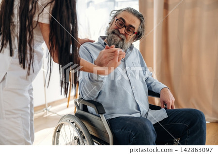 Elderly man in wheelchair interacts with caregiver in white uniform, bright indoor setting with natural light and soft curtains in the background Elderly man in wheelchair interacts with caregiver in white uniform, bright indoor setting with natural light and soft curtains in the background 142863097