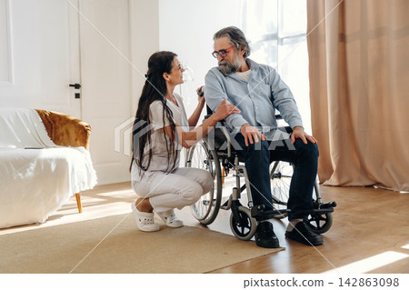 Female caregiver in white scrubs assists elderly man in wheelchair inside a bright living room with large windows and soft furnishings Female caregiver in white scrubs assists elderly man in wheelchair inside a bright living room with large windows and soft furnishings 142863098