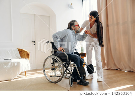 Elderly man in wheelchair assisted by caregiver in white uniform inside a bright, modern living room with wooden flooring and large windows Elderly man in wheelchair assisted by caregiver in white uniform inside a bright, modern living room with wooden flooring and large windows 142863104