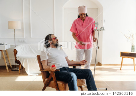 Female nurse in pink scrubs assists male patient with gray hair sitting in wooden chair, medical equipment visible in bright, modern room with soft lighting Female nurse in pink scrubs assists male patient with gray hair sitting in wooden chair, medical equipment visible in bright, modern room with soft lighting 142863116