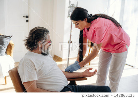 Female nurse in pink scrubs prepares to administer a medical procedure to an elderly male patient seated in a bright, modern healthcare environment Female nurse in pink scrubs prepares to administer a medical procedure to an elderly male patient seated in a bright, modern healthcare environment 142863123