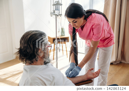 Female nurse in pink scrubs administers medical care to elderly male patient seated in a bright room with medical equipment and wooden furniture in the background Female nurse in pink scrubs administers medical care to elderly male patient seated in a bright room with medical equipment and wooden furniture in the background 142863124