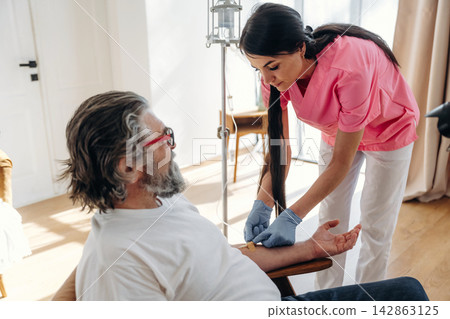 Female healthcare worker in pink scrubs prepares to administer a medical procedure to an elderly male patient seated in a well-lit room with wooden flooring Female healthcare worker in pink scrubs prepares to administer a medical procedure to an elderly male patient seated in a well-lit room with wooden flooring 142863125