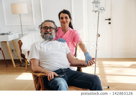 Female nurse in pink scrubs assists elderly man with gray beard seated in chair during medical procedure in bright, modern healthcare setting Female nurse in pink scrubs assists elderly man with gray beard seated in chair during medical procedure in bright, modern healthcare setting 142863126