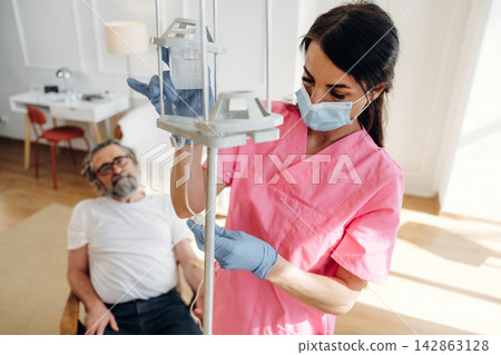 Female nurse in pink scrubs adjusts IV drip for elderly male patient seated in a bright hospital room with modern furniture and soft lighting Female nurse in pink scrubs adjusts IV drip for elderly male patient seated in a bright hospital room with modern furniture and soft lighting 142863128
