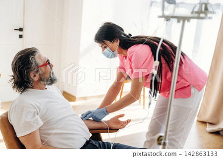 Healthcare professional in pink scrubs assists elderly male patient with intravenous therapy in a well-lit room with medical equipment and natural light 142863133