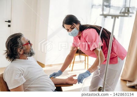 Female nurse in pink scrubs assists elderly male patient with intravenous therapy in a well-lit hospital room with wooden flooring and minimalistic decor Female nurse in pink scrubs assists elderly male patient with intravenous therapy in a well-lit hospital room with wooden flooring and minimalistic decor 142863134
