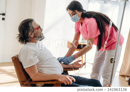 Female nurse in pink scrubs assists elderly male patient with intravenous therapy in a bright, modern healthcare setting with natural light streaming through windows Female nurse in pink scrubs assists elderly male patient with intravenous therapy in a bright, modern healthcare setting with natural light streaming through windows 142863136