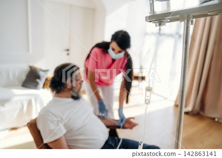Healthcare professional in pink scrubs administers IV treatment to male patient seated in chair at home, with medical equipment and bright interior visible in the background Healthcare professional in pink scrubs administers IV treatment to male patient seated in chair at home, with medical equipment and bright interior visible in the background 142863141