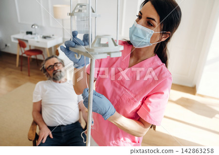 Female nurse in pink scrubs adjusts IV drip for elderly male patient seated in a modern room with wooden flooring and minimalistic furniture Female nurse in pink scrubs adjusts IV drip for elderly male patient seated in a modern room with wooden flooring and minimalistic furniture 142863258