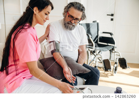 Female nurse in pink scrubs measures blood pressure of elderly man with gray beard sitting on a couch in a well-lit room with wheelchairs in the background Female nurse in pink scrubs measures blood pressure of elderly man with gray beard sitting on a couch in a well-lit room with wheelchairs in the background 142863260