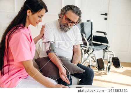 Female healthcare worker in pink scrubs measures blood pressure of elderly man in white shirt, with wheelchairs visible in a bright indoor setting 142863261