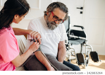 Healthcare professional in pink scrubs measures blood pressure of elderly man with gray beard sitting on couch in home setting with wheelchair visible in the background Healthcare professional in pink scrubs measures blood pressure of elderly man with gray beard sitting on couch in home setting with wheelchair visible in the background 142863264