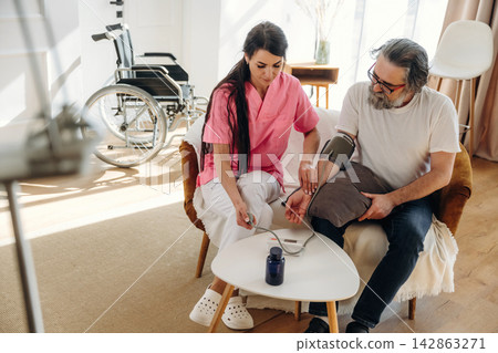 Female nurse in pink scrubs measures blood pressure of senior man in a cozy living room with a wheelchair and modern furniture in the background Female nurse in pink scrubs measures blood pressure of senior man in a cozy living room with a wheelchair and modern furniture in the background 142863271