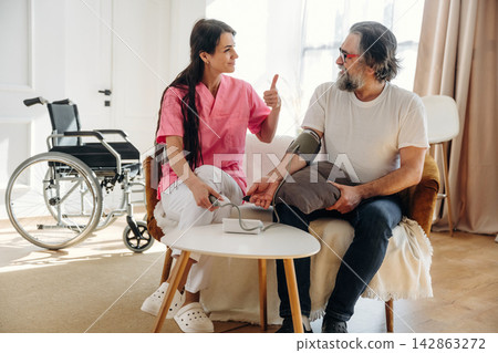 Female healthcare professional in pink scrubs checks blood pressure of elderly man in white t-shirt seated on a chair with wheelchair visible in a bright indoor setting Female healthcare professional in pink scrubs checks blood pressure of elderly man in white t-shirt seated on a chair with wheelchair visible in a bright indoor setting 142863272