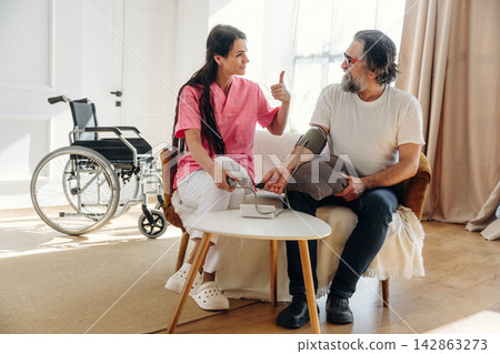 Female healthcare worker in pink scrubs checks blood pressure of male patient sitting on a couch in a well-lit room with a wheelchair nearby Female healthcare worker in pink scrubs checks blood pressure of male patient sitting on a couch in a well-lit room with a wheelchair nearby 142863273