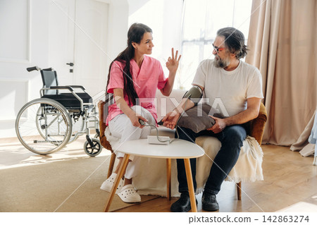 Female healthcare worker in pink scrubs checks blood pressure of elderly man sitting in a chair with a wheelchair visible in a bright, modern indoor setting Female healthcare worker in pink scrubs checks blood pressure of elderly man sitting in a chair with a wheelchair visible in a bright, modern indoor setting 142863274