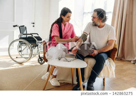 Female nurse in pink scrubs measures blood pressure of elderly man in white shirt seated on chair with wheelchair visible in a well-lit indoor setting Female nurse in pink scrubs measures blood pressure of elderly man in white shirt seated on chair with wheelchair visible in a well-lit indoor setting 142863275