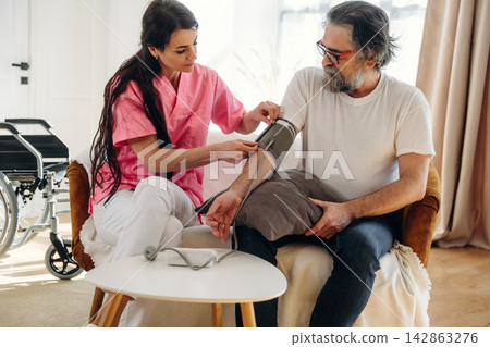 Female nurse in pink scrubs measures blood pressure of elderly man in white t-shirt while seated in a cozy living room with wheelchair in the background Female nurse in pink scrubs measures blood pressure of elderly man in white t-shirt while seated in a cozy living room with wheelchair in the background 142863276