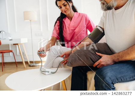 Female healthcare professional in pink scrubs measures blood pressure of senior man seated on couch in modern living room with light-colored walls and furniture 142863277