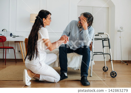 Female healthcare worker in white uniform kneels beside elderly man with gray hair, discussing care in a bright room with a walker and furniture in the background Female healthcare worker in white uniform kneels beside elderly man with gray hair, discussing care in a bright room with a walker and furniture in the background 142863279