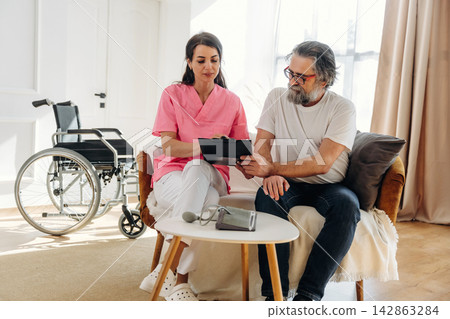 Female caregiver in pink scrubs assists elderly man with tablet in a cozy living room, wheelchair positioned nearby, sunlight streaming through large windows Female caregiver in pink scrubs assists elderly man with tablet in a cozy living room, wheelchair positioned nearby, sunlight streaming through large windows 142863284