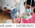 Female nurse in pink scrubs adjusts IV drip with clear fluid in hospital room, medical equipment and patient bed visible in the background 142863622