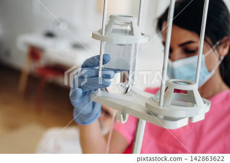 Female nurse in pink scrubs adjusts IV drip with clear fluid in hospital room, medical equipment and patient bed visible in the background Female nurse in pink scrubs adjusts IV drip with clear fluid in hospital room, medical equipment and patient bed visible in the background 142863622