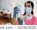 Female healthcare worker in pink scrubs adjusts IV drip stand while wearing gloves and a mask in a bright hospital room with patient in the background 142863626