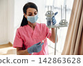 Female healthcare worker in pink scrubs adjusts IV drip stand while wearing gloves and a mask in a well-lit medical facility with large windows 142863628