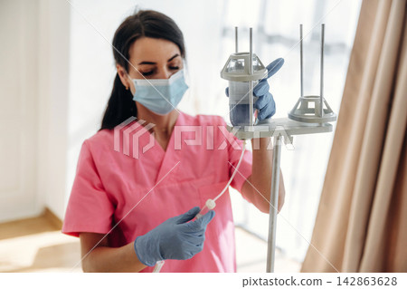 Female healthcare worker in pink scrubs adjusts IV drip stand while wearing gloves and a mask in a well-lit medical facility with large windows Female healthcare worker in pink scrubs adjusts IV drip stand while wearing gloves and a mask in a well-lit medical facility with large windows 142863628
