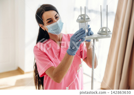 Female healthcare worker in pink scrubs adjusts IV drip stand while wearing a mask and gloves, bright hospital room with natural light and curtains in the background 142863630
