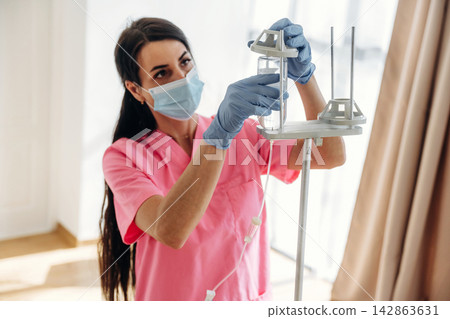 Female healthcare worker in pink scrubs adjusts IV drip in a bright hospital room with large windows and soft natural light illuminating the space Female healthcare worker in pink scrubs adjusts IV drip in a bright hospital room with large windows and soft natural light illuminating the space 142863631