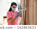 Female healthcare worker in pink scrubs adjusts medical equipment while wearing a mask and gloves, with soft lighting and curtains in a hospital or clinic setting 142863633