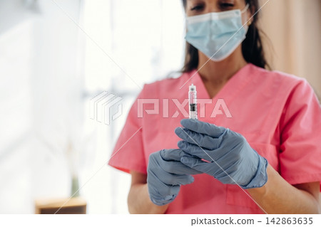 Healthcare professional in pink scrubs holds a syringe filled with insulin, wearing blue gloves and a face mask, in a well-lit clinical environment with soft natural light Healthcare professional in pink scrubs holds a syringe filled with insulin, wearing blue gloves and a face mask, in a well-lit clinical environment with soft natural light 142863635