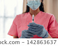 Healthcare professional in pink scrubs holds a syringe with insulin, wearing blue gloves and a face mask, in a well-lit medical environment with soft background 142863637