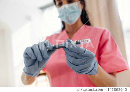 Healthcare professional in pink scrubs prepares insulin syringe for diabetes treatment, wearing blue gloves and a surgical mask in a clinical setting with natural light Healthcare professional in pink scrubs prepares insulin syringe for diabetes treatment, wearing blue gloves and a surgical mask in a clinical setting with natural light 142863640