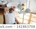 Elderly man with gray beard sitting in a chair, holding his forehead in discomfort, with an IV drip stand and medical equipment in a bright hospital room 142863708