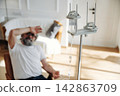 Elderly man with gray beard sitting in chair, resting hand on forehead, IV drip stand beside him in bright room with soft furnishings and natural light 142863709