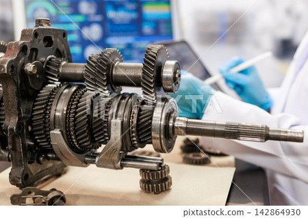 Close-up of a manual transmission gearbox with exposed metal gears on a workbench, while a mechanic in gloves inspects parts in a bright repair workshop. Close-up of a manual transmission gearbox with exposed metal gears on a workbench, while a mechanic in gloves inspects parts in a bright repair workshop. 142864930